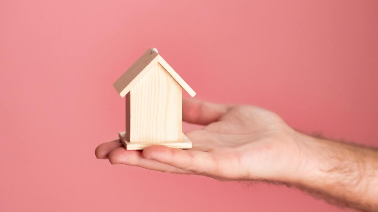 Main tenant un petit modèle de maison en bois sur un fond rose doux, symbolisant l'immobilier et l'investissement immobilier.