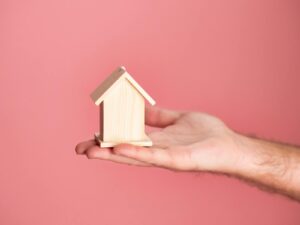 Main tenant un petit modèle de maison en bois sur un fond rose doux, symbolisant l'immobilier et l'investissement immobilier.