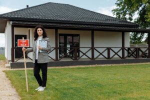 Agent immobilier debout devant une maison avec un panneau « Maison à vendre » sur une pelouse herbeuse.