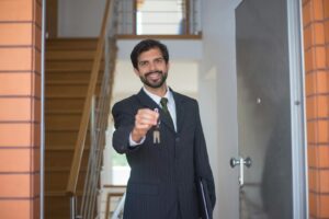 Agent immobilier souriant en costume noir remettant des clés à travers une porte ouverte à l'intérieur d'une maison moderne.
