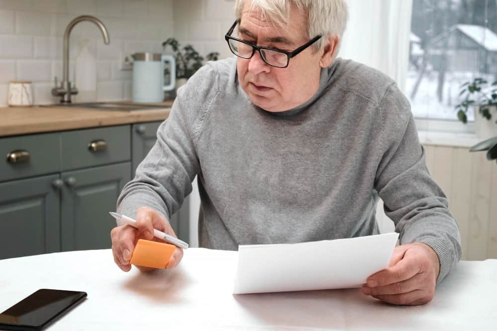 Un homme senior assis dans une cuisine regarde des documents financiers concernant le calcul de sa pension alimentaire.